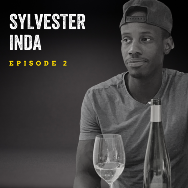 Black and white image of a man wearing a backwards ball cap and t-shirt looks to the left of the camera, with a glass of white wine and bottle on a table in front of him. He is in front of a dark background with text overlay reading "Sylvester Inda Episode 2."