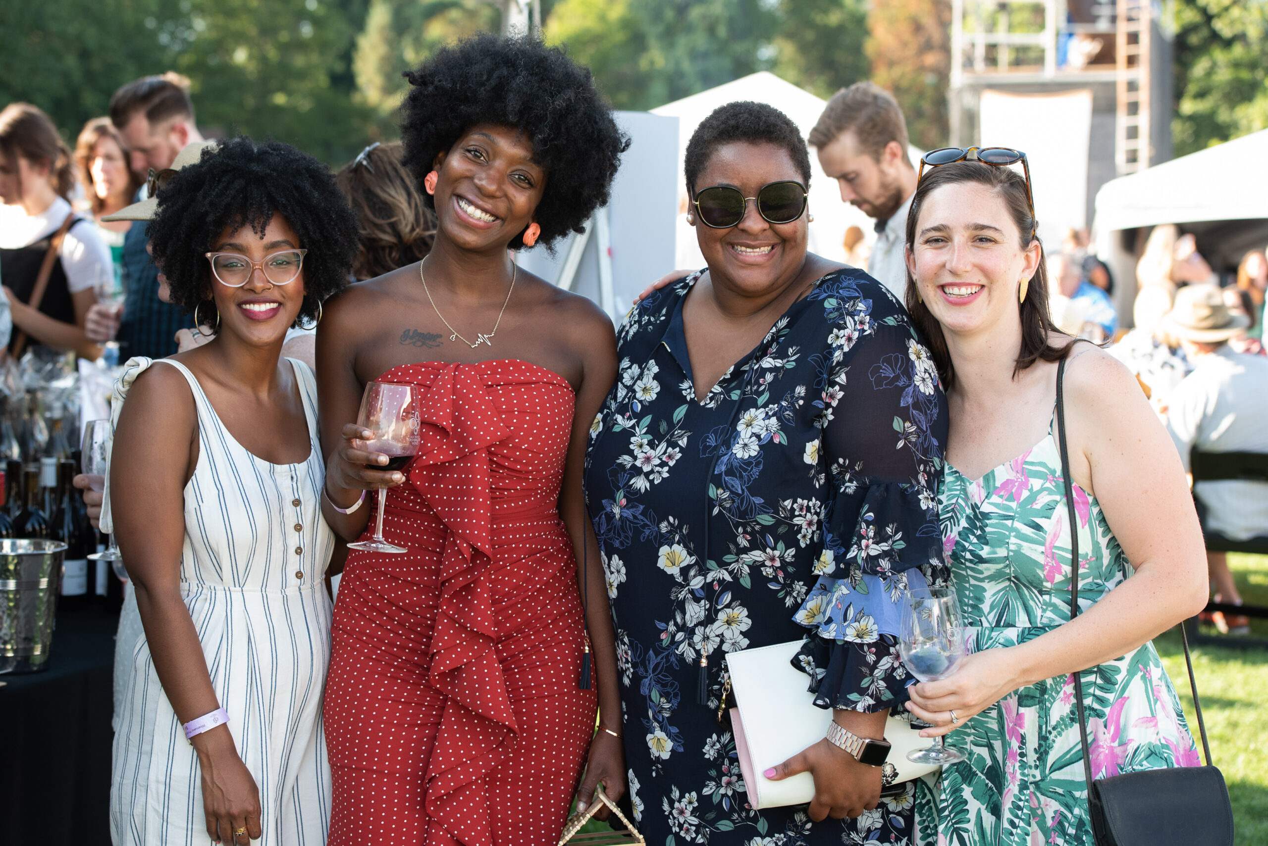 Four women stand together and smile. Two of them hold glasses of wine and they are wearing festive summer dresses. There are people and tents behind them.