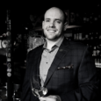 A black and white image of a man (Nick Davis) with short buzzed hair wearing a jacket and collared shirt smiles softly and holds a glass of white wine. The background is a dark room with bottles and a bar.