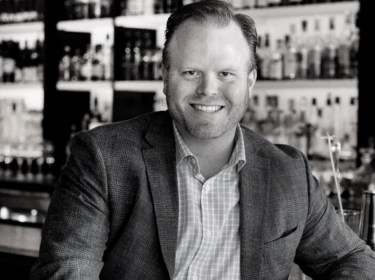 Black and white image of a man seated with his elbow on a bar. He wears a suit jacket and gingham button up shirt and smiles. Shelves of bottles are behind him.