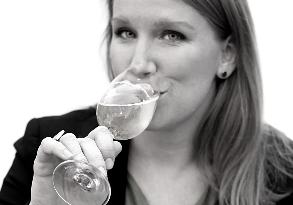A young woman sips from a glass of white wine. She has long medium dark hair and stud earrings in front of a light background.