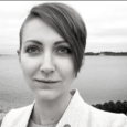 Black and white close-up image of a woman with a side-swept pixie haircut. She smiles wryly and wears a white collared blazer. A large body of water and distant shore are behind her.