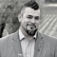 Black and white image of man with dark hair slicked up in a faux-hawk and a dark beard, wearing a grey jacket and collared shirt. There are rows of vines in the background.