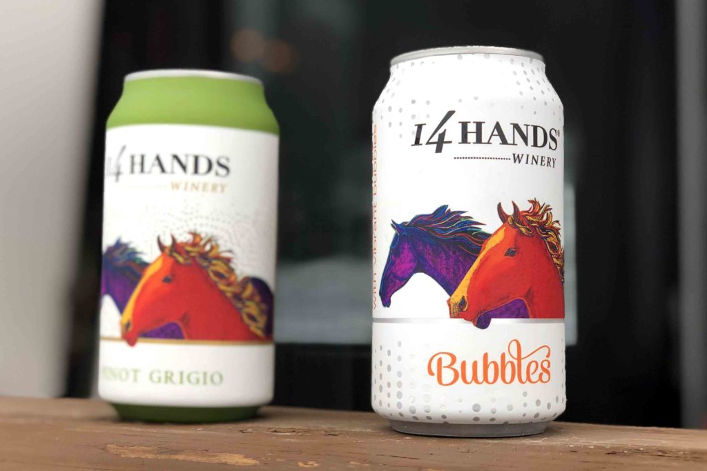 Two cans of wine on a brown table with white labels with "14 Hands" and red and purple horses.