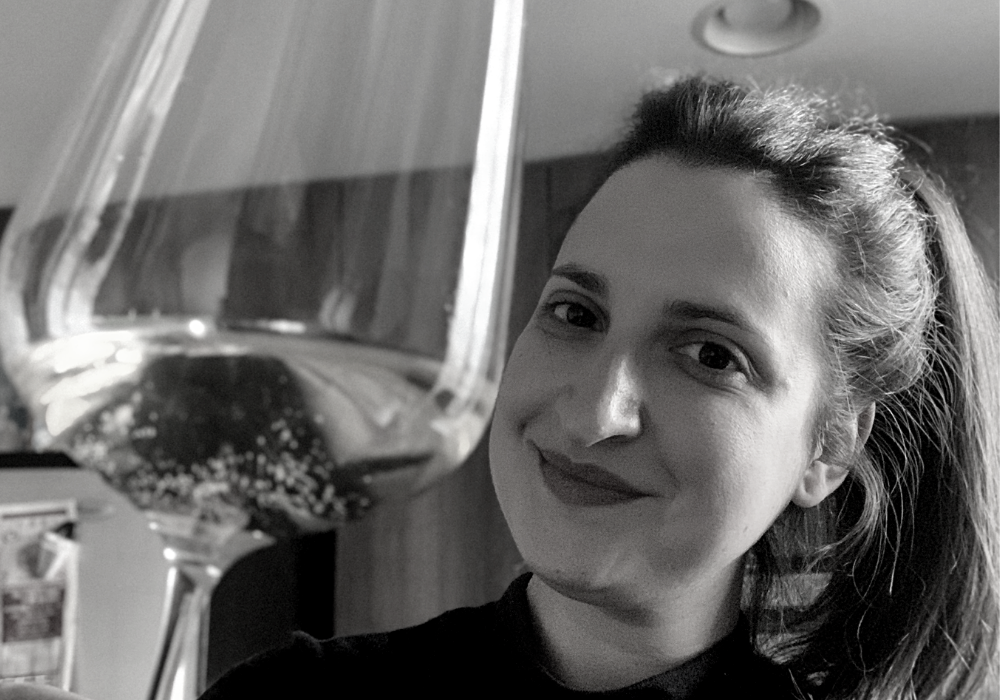 Black and white image of a woman with dark lipstick and long dark hair tied up, holding a glass of white wine to the camera. She is indoors with a dark wall behind her.
