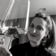 Black and white image of a woman with dark lipstick and long dark hair tied up, holding a glass of white wine to the camera. She is indoors with a dark wall behind her.