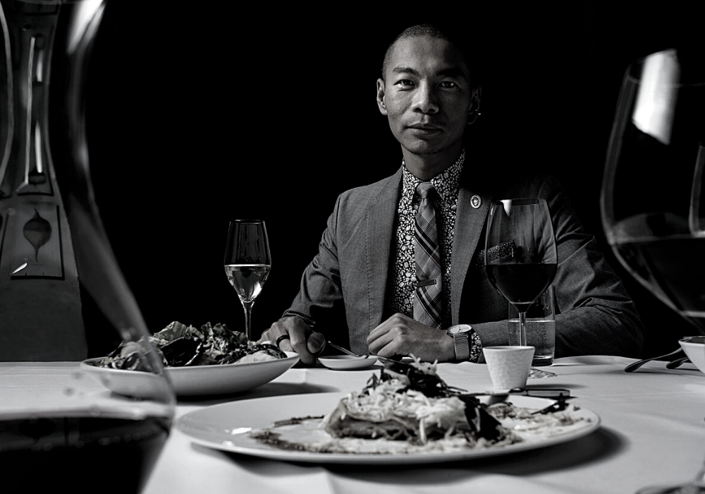 Black and white image of man wearing a dark suit, floral shirt, and plaid tie, seated at a table with a white table cloth, plates of food, and glasses of wine. He sits in front of a dark background.