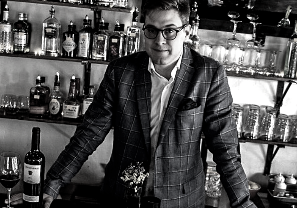 Black and white image of man leaning on a bar wearing a plaid jacket and dark glasses. There are shelves of bottles behind him.