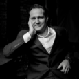 Black and white image of a seated man with his hand resting on his face. The man wears a black suit jacket and white button up shirt and smiles softly.