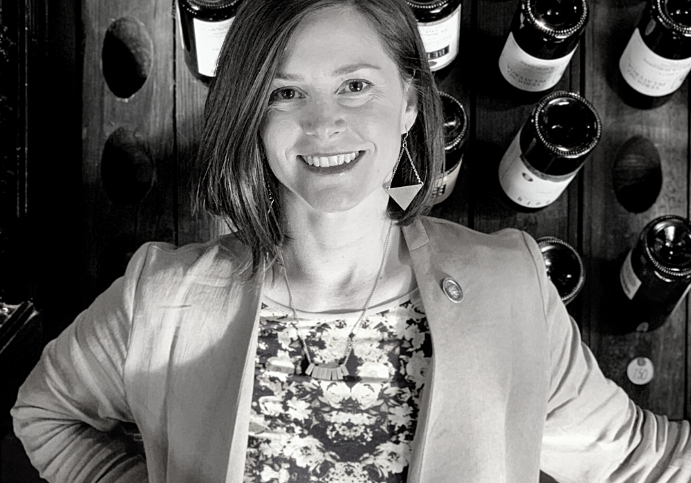 Black and white image of woman standing with hands on her hips, wearing a light suit jacket and floral shirt. A rack of wine bottles is behind her.
