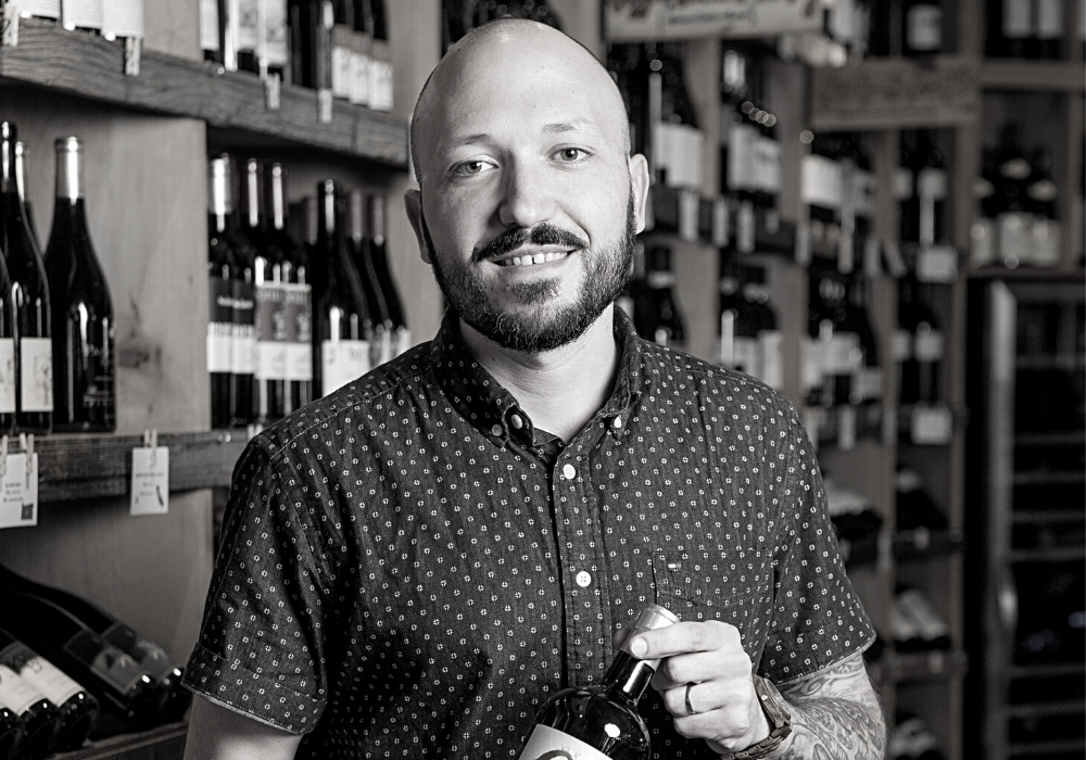 Black and white image of bald man with a dark beard holding a bottle of wine with both hands. The background is shelves of wine bottles.
