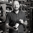 Black and white image of bald man with a dark beard holding a bottle of wine with both hands. The background is shelves of wine bottles.