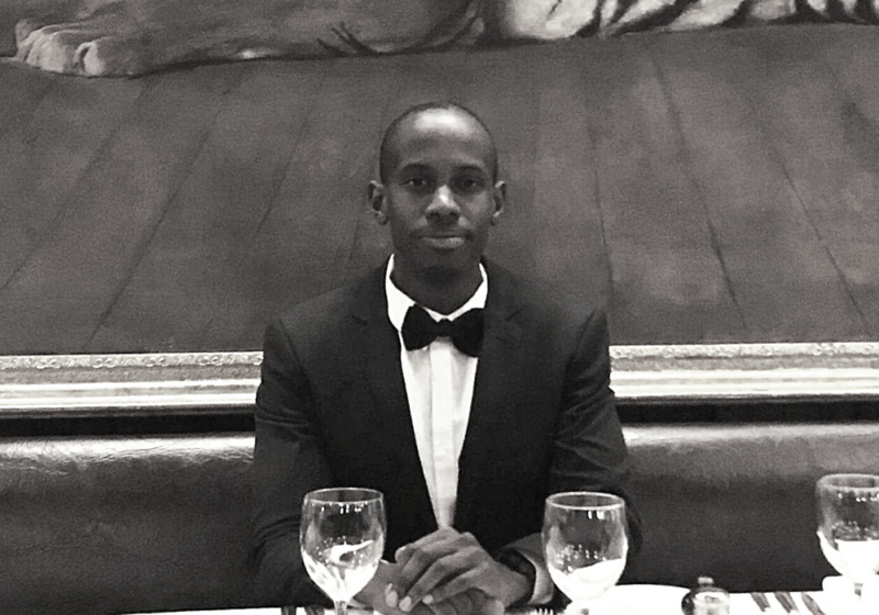A man wearing a dark suit jacket and bowtie sits at a restaurant booth with wine glasses in front of him.