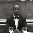 A man wearing a dark suit jacket and bowtie sits at a restaurant booth with wine glasses in front of him.