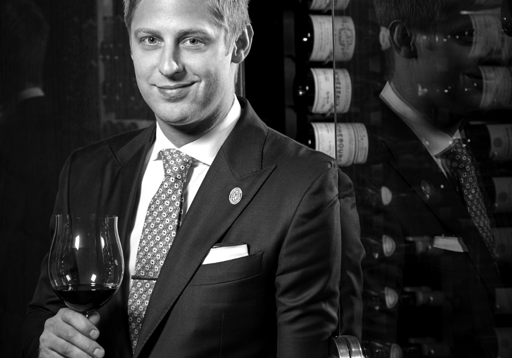 A man with scruffy blond hair wearing a suite and tie smiles and holds a glass of red wine in front of racks of wine bottles