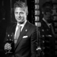 A man with scruffy blond hair wearing a suite and tie smiles and holds a glass of red wine in front of racks of wine bottles