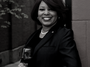 Black and white image of a woman with chin-length dark hair and wearing a black suit jacket leans against a stone wall outdoors with a glass of wine in her hand.