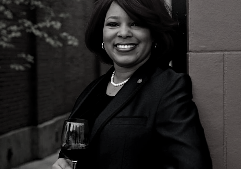 Black and white image of a woman with chin-length dark hair and wearing a black suit jacket leans against a stone wall outdoors with a glass of wine in her hand.