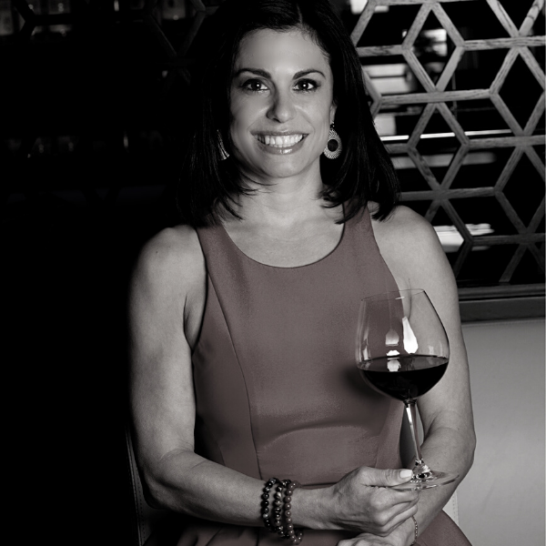 Black and white image of a woman seated and holding a glass of red wine. She has dark medium length hair and a bright smile, and wears a cocktail dress. She sits in front of a wine rack with geometric shapes.