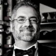 Black and white image of a man with grey and white hair and beard wearing a black suit and bowtie, smiling. He stands in front of shelves with bar paraphernalia.