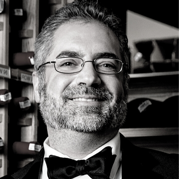 Black and white image of a man with grey and white hair and beard wearing a black suit and bowtie, smiling. He stands in front of shelves with bar paraphernalia.