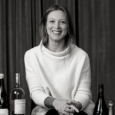 Black and white image of a young woman with light hair smiles broadly and wears a loose white sweater. She is seated in front of a velvet curtain with several bottles on a table around her.