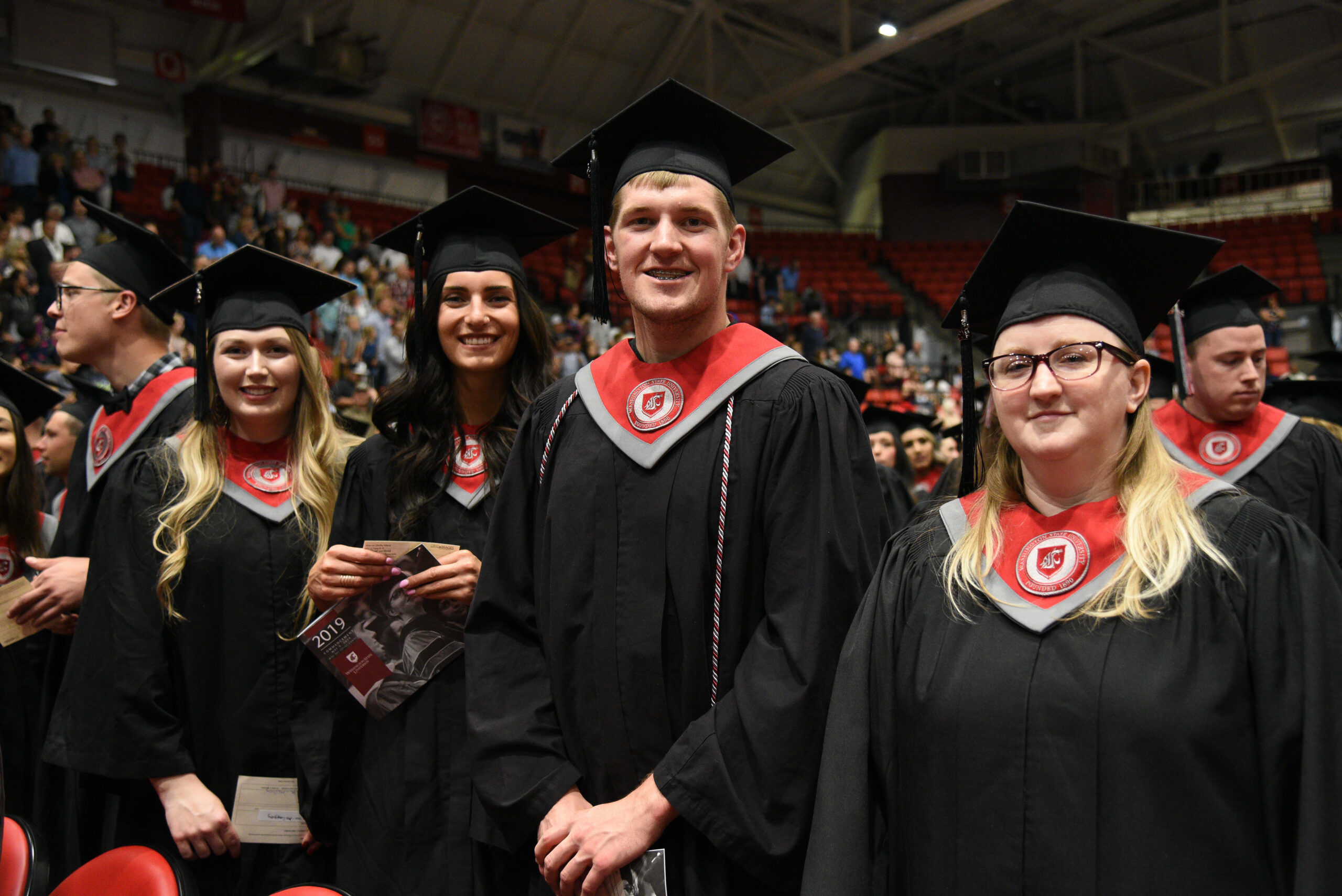 Four people in black graduation robes, black graduation hats, and red collars smile at the camera in front of a crowded auditorium.