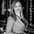 A woman with long blond-brown hair with dark lipstick smiles and holds a glass of red wine. She stands in front of racks of wine bottles and wears a light colored jacket..