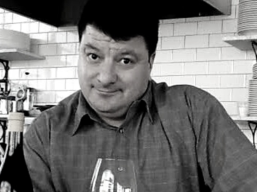 black and white image of a man (Lenny Rede) with short dark hair holds a glass of red wine resting on a counter. A wall of white subway tiles are behind him.