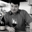 black and white image of a man (Lenny Rede) with short dark hair holds a glass of red wine resting on a counter next to a bottle of wine. White subway tiles and shelves with plates are behind him.