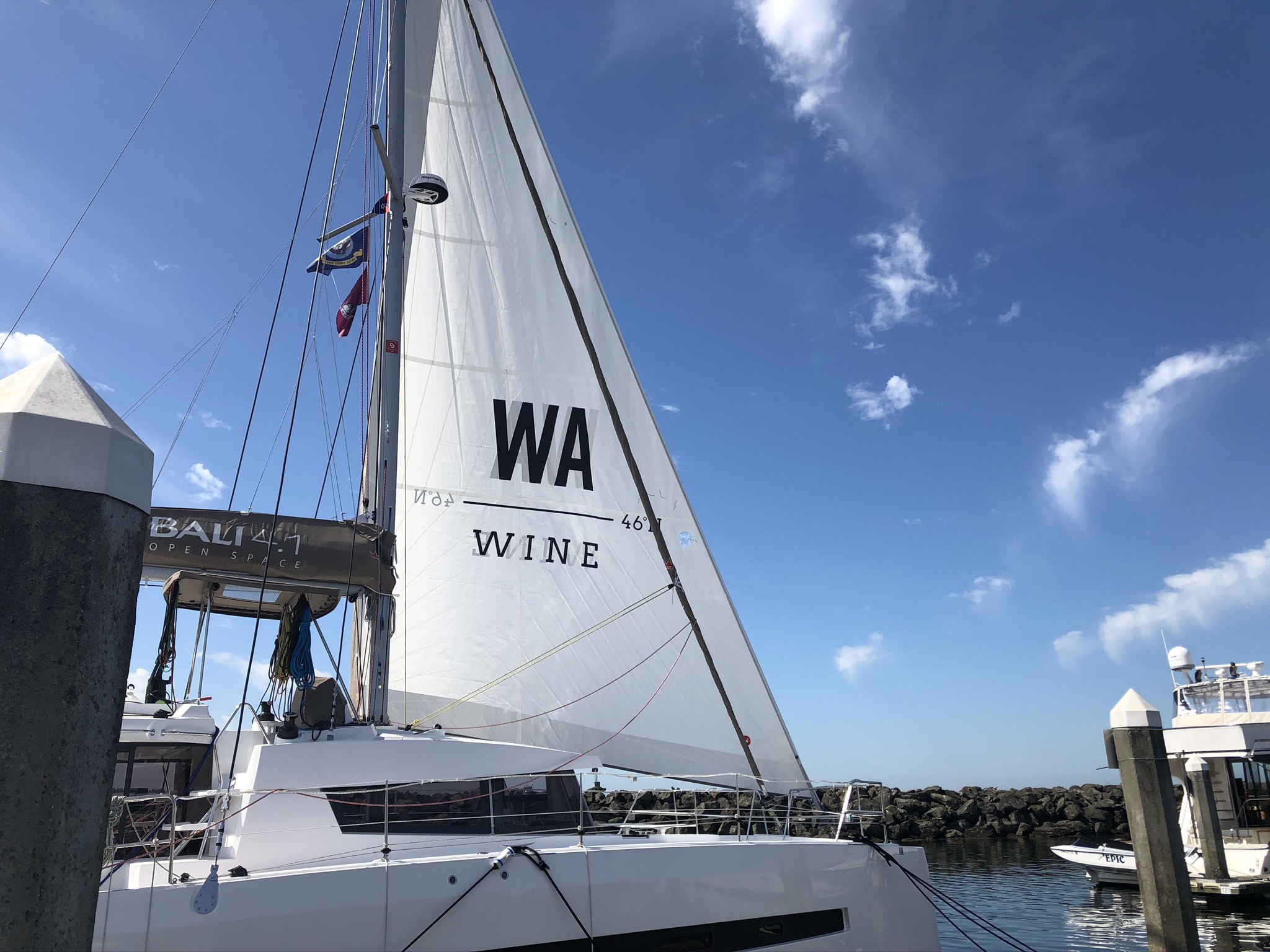 A white sail with a black WA Wine logo on a white sail boat, in front of a bright blue sky.