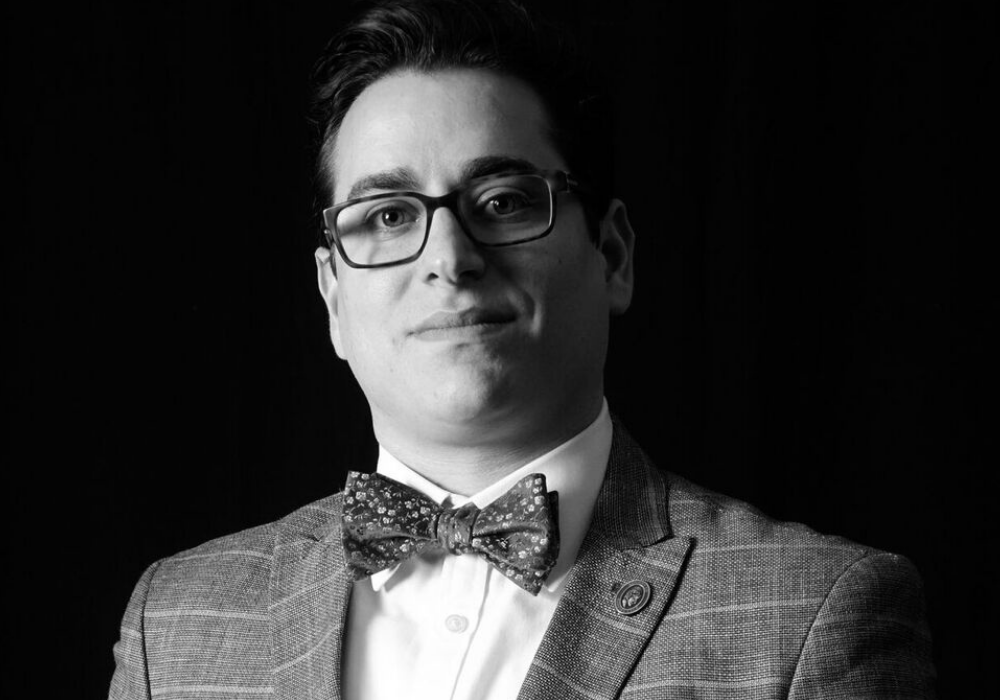 Black and white image of a man wearing a plaid jacket and bowtie, with glasses and short dark hair. He is in front of a dark background.