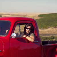 Woman leans out the driver's seat window of a vintage red pick-up truck. Green vines and tan fields are in the background.