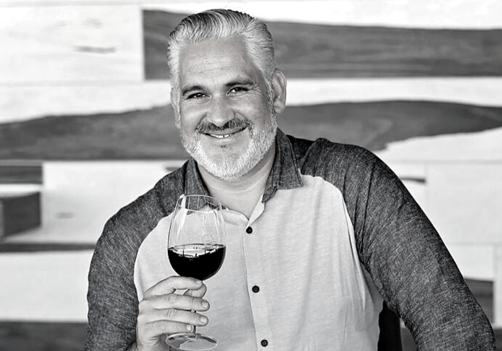 Black and white image of a man with grey and white hair and short beard, smiling and holding a glass of red wine. He wears a shirt with dark sleeves and white front with buttons.