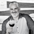 Black and white image of a man with grey and white hair and short beard, smiling and holding a glass of red wine. He wears a shirt with dark sleeves and white front with buttons.