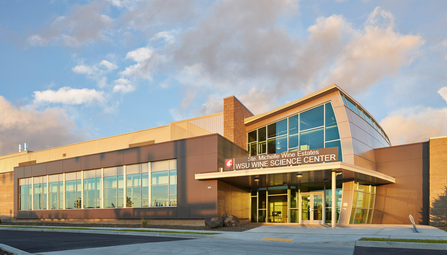 A large brown and tan academic building with a sign above the door that reads "Ste. Michelle Wine Estates WSU Wine Science Center" under a blue sky with wispy grey clouds.