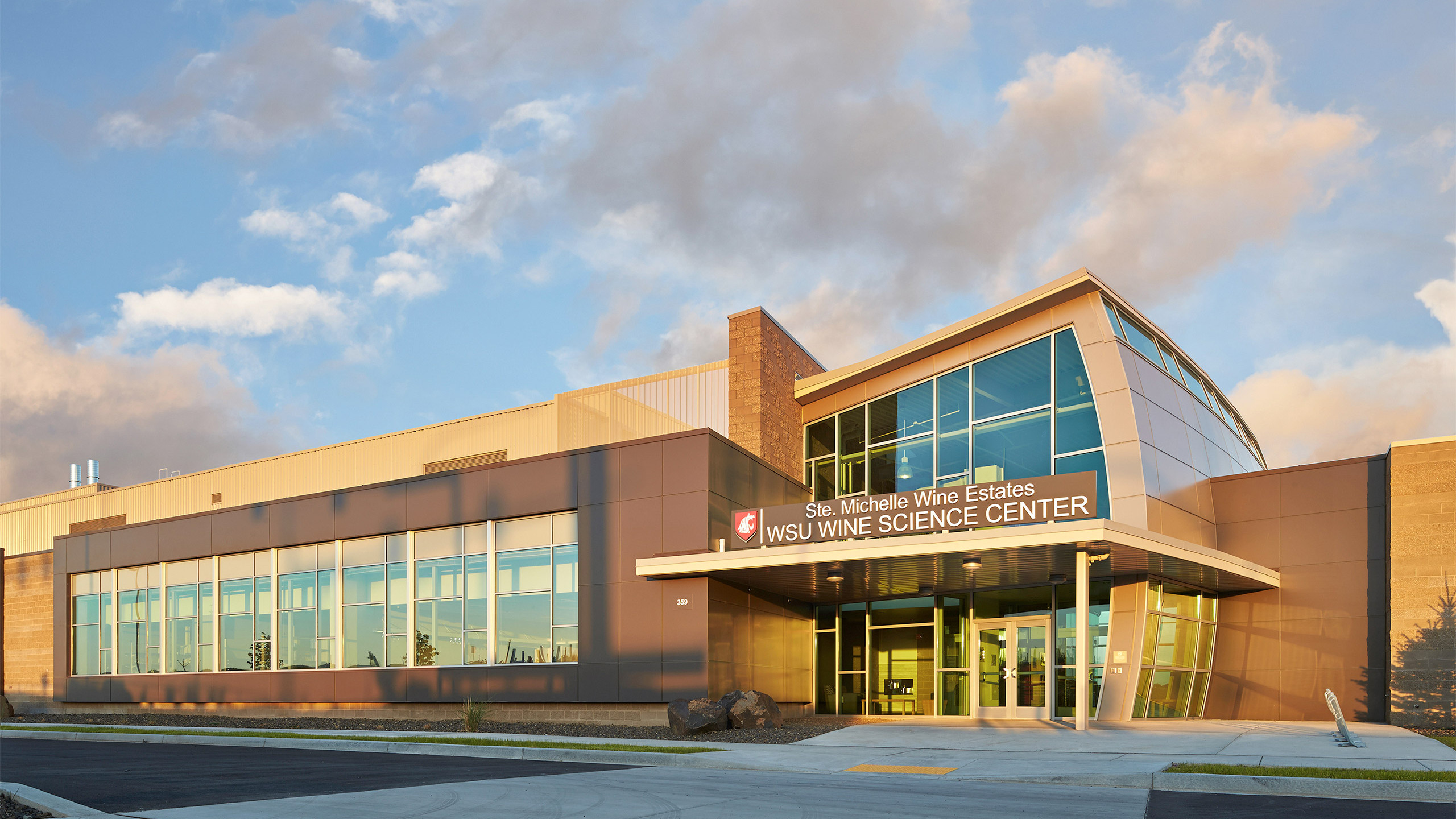 A large brown and tan academic building with a sign above the door that reads "Ste. Michelle Wine Estates WSU Wine Science Center" under a blue sky with wispy grey clouds.