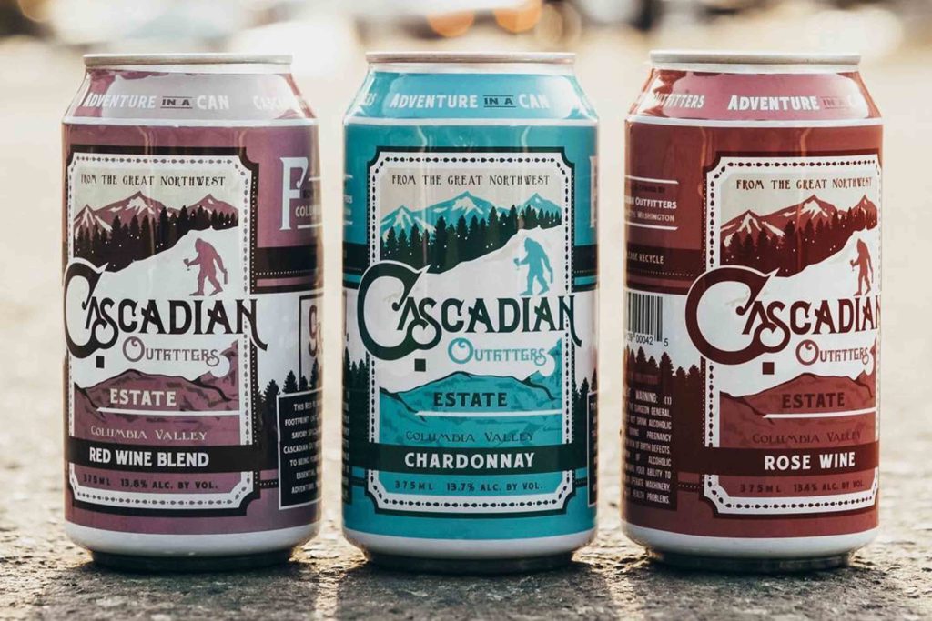 Three cans of Cascadian Outfitters wine, in purple, blue, and maroon labels. They are outside, on concrete.