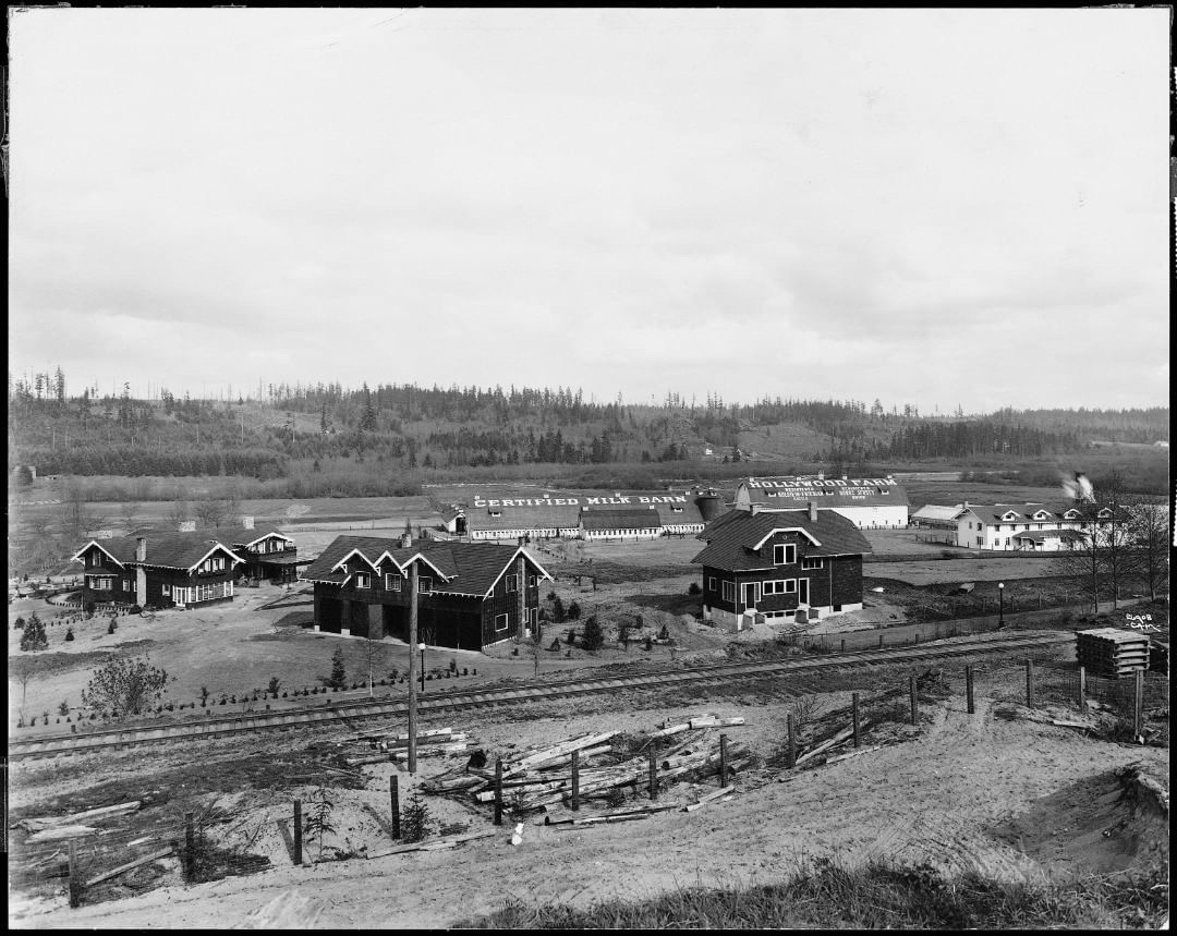 black and white image of dark buildings with white trim, white barn and agriculture buildings, and a railroad track and post fence in the foreground. 