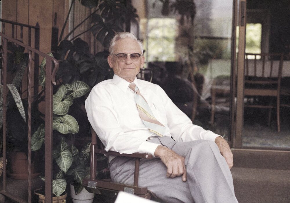 An older man in a white shirt and striped tie and slacks smiles softly and sits in a chair in a sunroom in front of house plants.