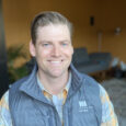 A man with brown hair combed back in a blue vest and orange and blue plaid shirt smiles in front of a couch and orange wall.