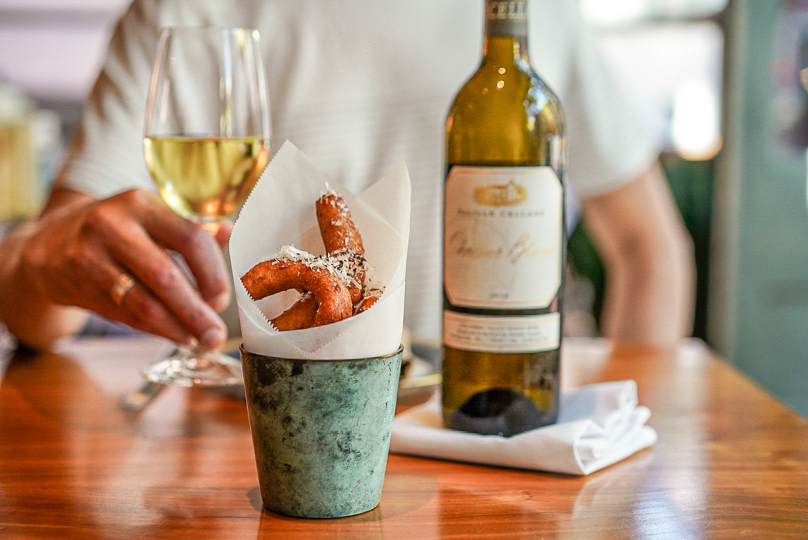 A wooden table with a metal cup lined with parchment paper and holding onion rings, a bottle of wine, and a glass of white wine. A hand holds the glass and a torso is out of focus behind the table.