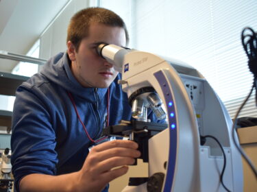 A young man with short-cropped hair wearing a blue sweatshirt looks into a microscope.