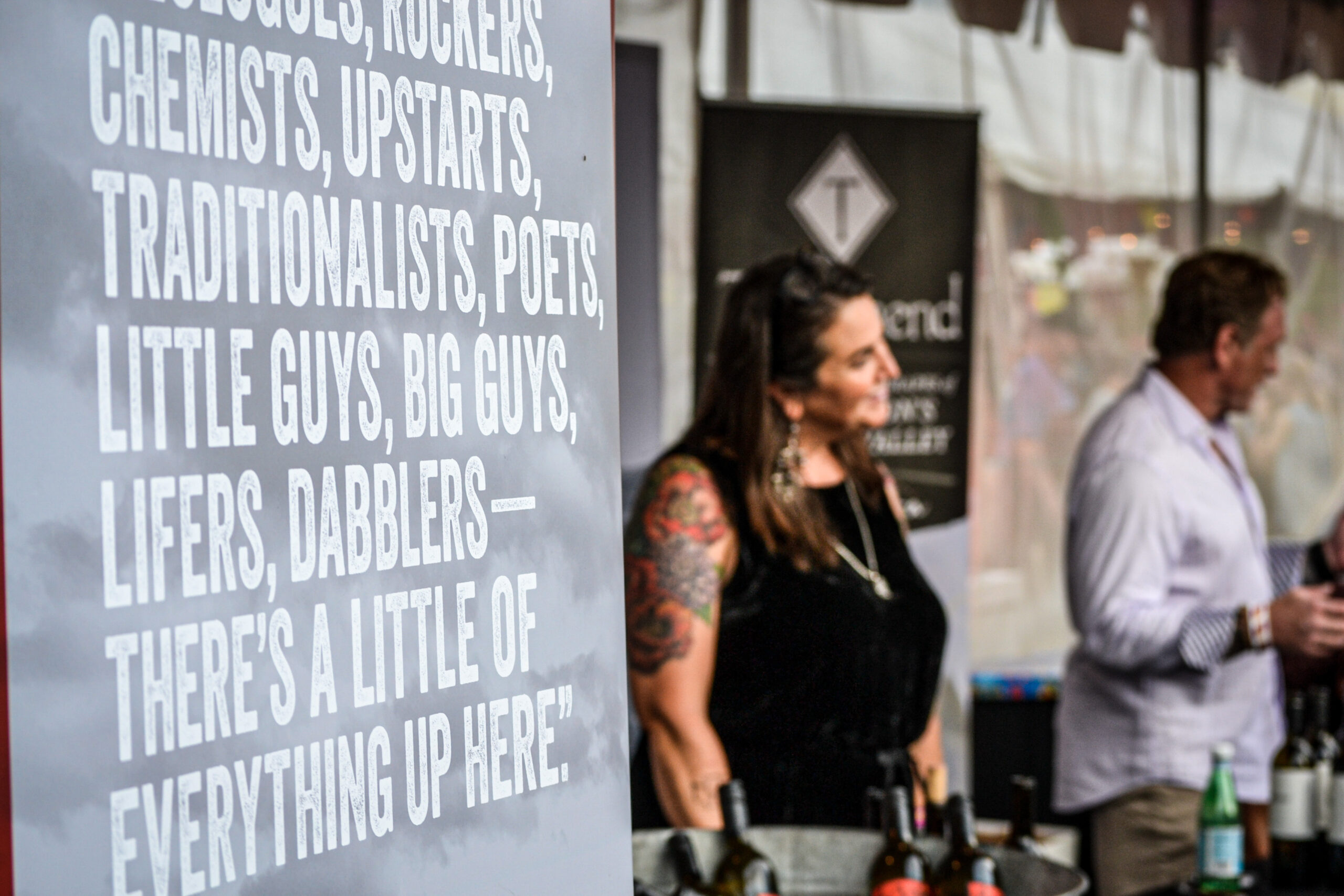 A banner with white lettering with a woman in a black shirt and tattooed arm smiling and a man next to her in the background.