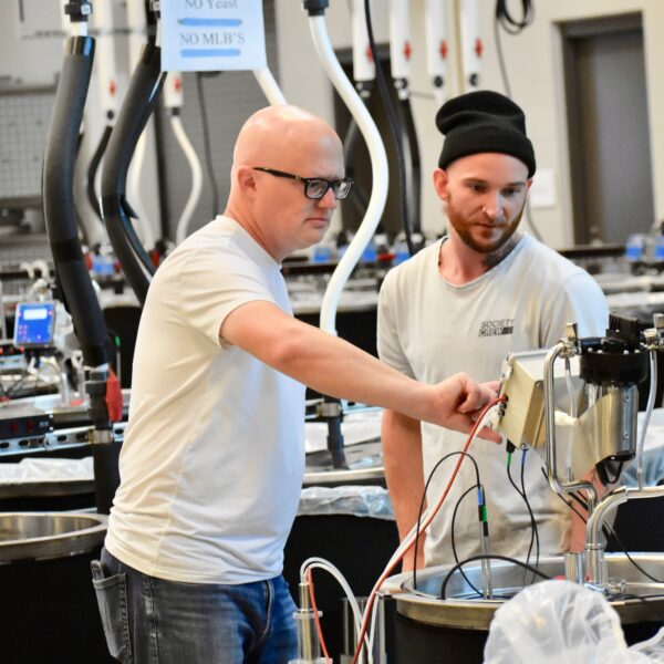 A bald man with glasses touches a piece of equipment in a science lab. A young man in a black beanie stands next to him and watches.
