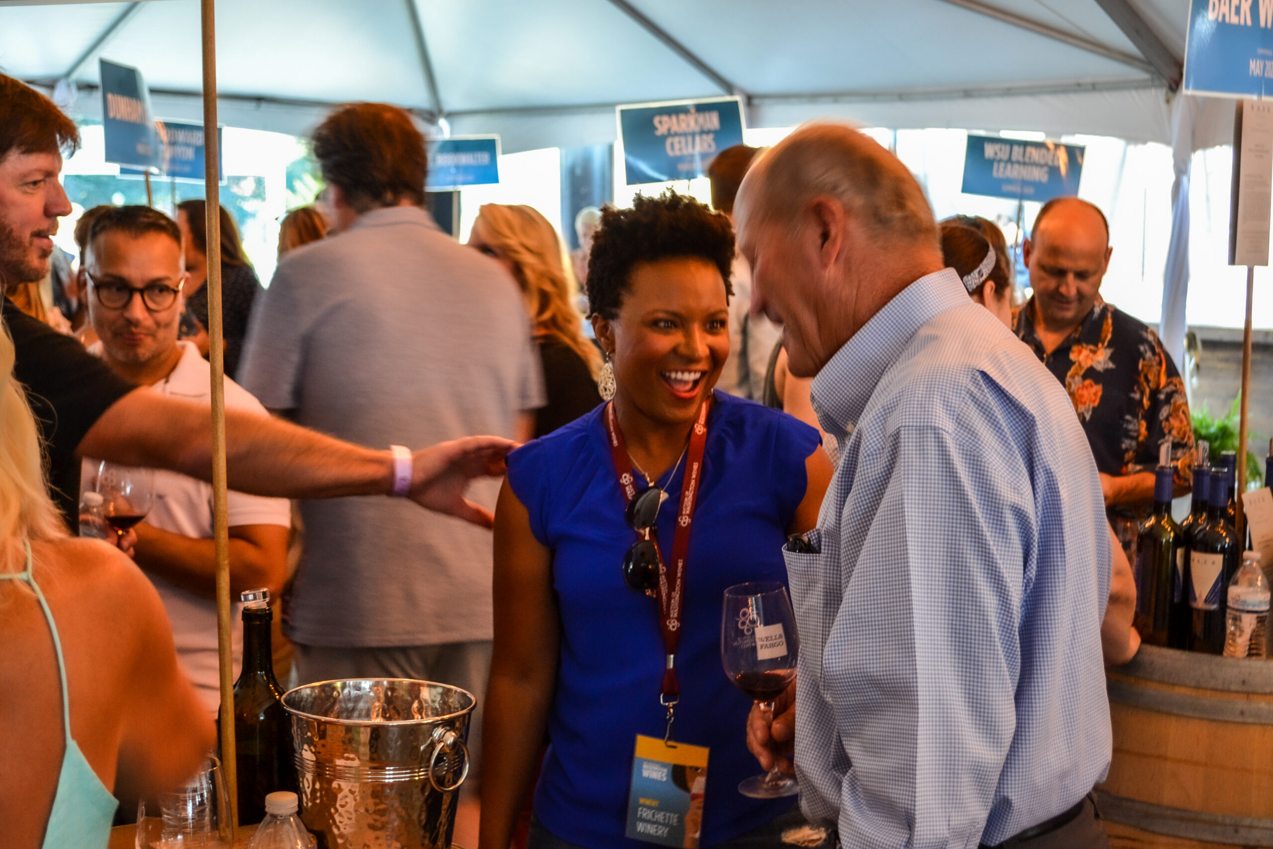 A woman with dark hair in a blue shirt talks with a man in light blue button-up shirt in a crowd of people under a tent.