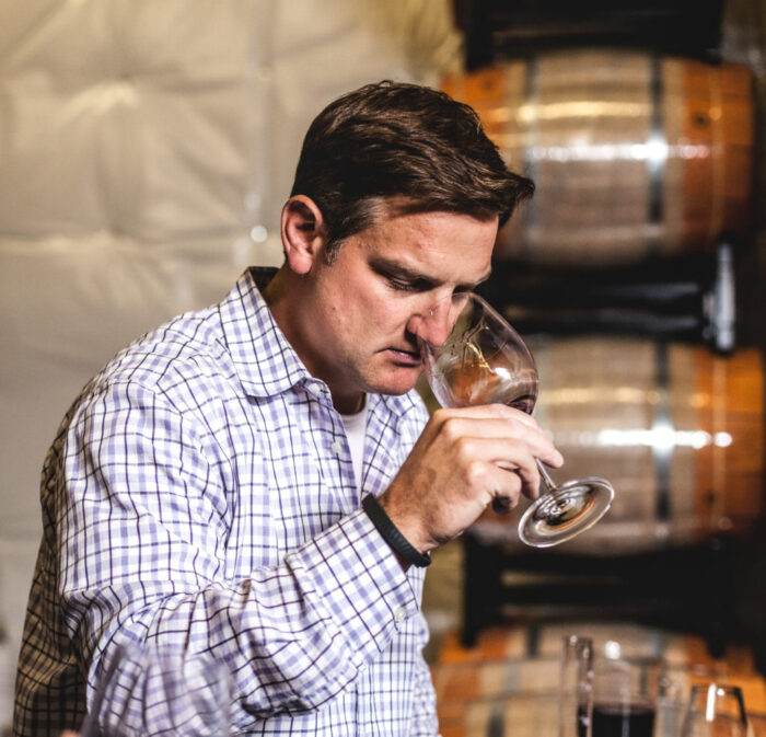 A man in a purple and white plaid shirt holds a glass of wine to his nose. Barrels are in the background.