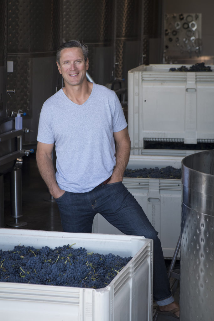 A man in a blue t-shirt smiles and stands between bins of indigo wine grapes.