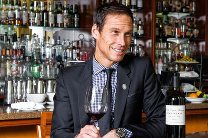 A man smiles while sitting in front of a bar with many bottles. He has a glass of red wine in his hand resting on the table and a bottle of wine in front of him.