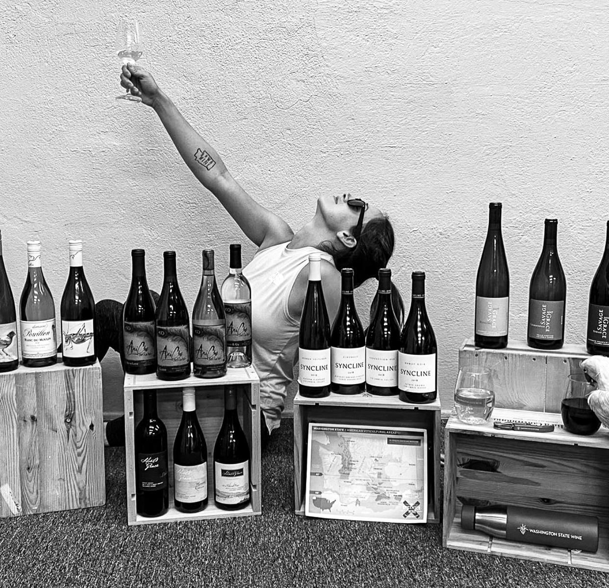 Black and white image of a woman holding a hand with a glass of wine in the air with her head back. She is behind wooden boxes with bottles of wine lined up on them.